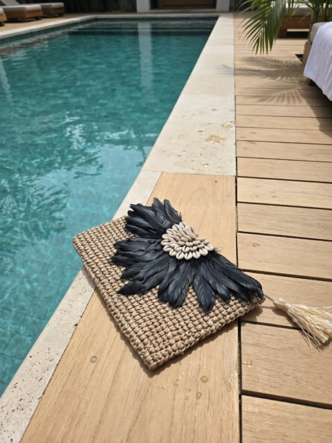 A woven handmade raffia clutch bag decorated with black feathers and white shells, resting on a wooden deck next to a swimming pool.