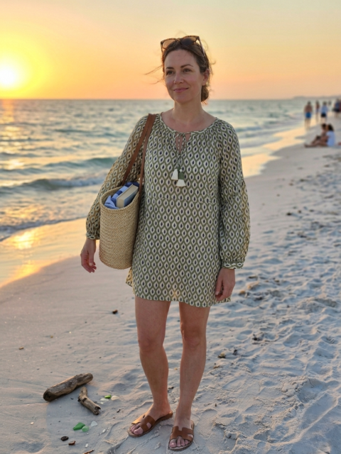 A woman standing on a sandy beach at sunset wearing a green and white geometric block print cotton top with tassels, carrying a straw bag.